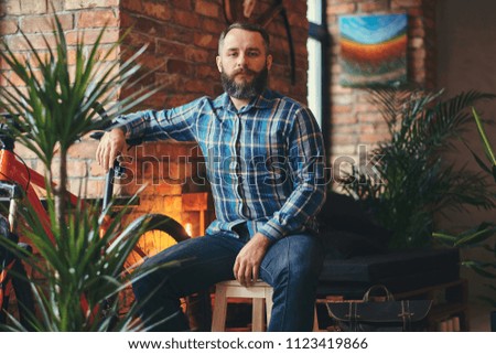 Handsome bearded hipster male in a blue fleece shirt and jeans sitting on a wooden stool at a studio with a loft interior.