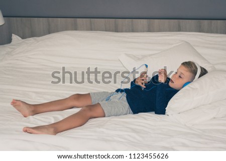 Small kid with headphones using smart phone and watching cartoons while lying down on bed. 