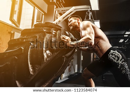 Muscular bearded tattooed fitness shirtless man moving large tire in gym. Concept lifting, workout cross fit training. Royalty-Free Stock Photo #1123584074