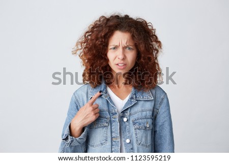 Closeup of confused displeased lovely young woman with red curly hair in denim jacket asking quetion and pointing at herself with finger isolated over white background