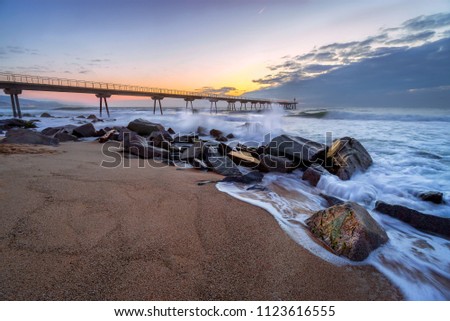Landscape photography of rocks barrier holding the waves and a long petroleum bridge at the beach at sunrise with a stormy and rough sea with lots of waves