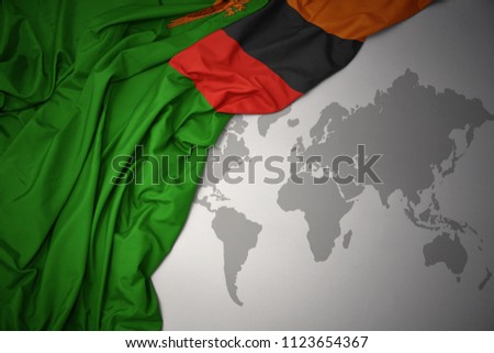 waving colorful national flag of zambia on a gray world map background.