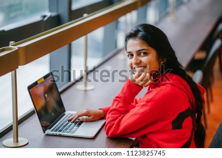 A portrait of a young and attractive Indian Asian student woman girl sitting at a wooden desk and working on her laptop computer during the day. She is pretty and is smiling as she looks at the camera