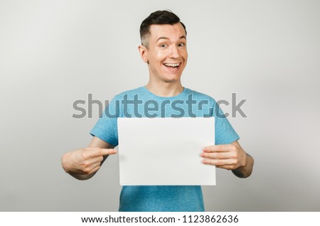 Young smiling guy dressed in a blue t-shirt holds a sheet of paper of A4    on a light background.
