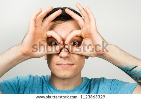 Young guy dressed in a blue t-shirt looks through binoculars from the palms of his hands  on a light background. Close-up portrait