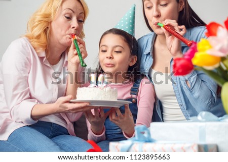 Two women and girl together at home celebrating birthday sitting on sofa wearing festive caps girl holding cake blowing candles making wish while mom and granny blowing party horns close-up.