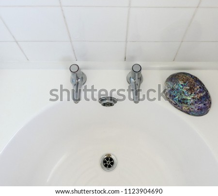 Partial view of empty white bath and tiles looking down on chromed taps, overflow and plug hole. Iridescent colour provided by Abalone shell, with reflections of taps and shell. Royalty-Free Stock Photo #1123904690