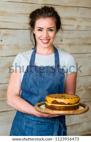 a young woman stands proudly in front of a white rustic wooden wall of a cafe showing off her fresh cake.