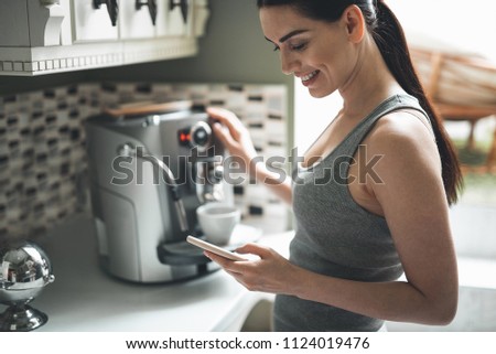 Side view of smiling attractive woman standing by coffee machine with smartphone in hands. She is chatting online with delight Royalty-Free Stock Photo #1124019476