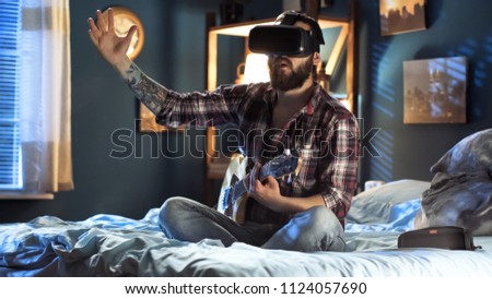 Casual man wearing VR goggles and give virtual concert with guitar while sitting on bed at home at night Royalty-Free Stock Photo #1124057690