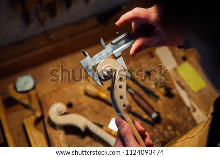 Master artisan luthier working on creation of a violin scroll. detailed work on wood with tools. Men's hands Measures the thickness of the scroll with meter . on the background of a workshop Royalty-Free Stock Photo #1124093474
