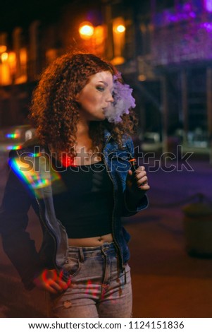 Young attractive red-haired girl smokes electronic cigarette. She walks down street. Evening city lights and signs flashing.