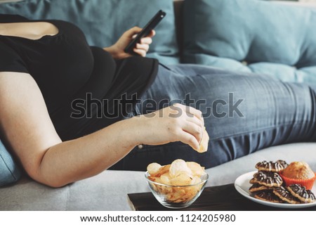 g, sedentary lifestyle, compulsive overeating. Obese woman laying on sofa with smartphone eating chips Royalty-Free Stock Photo #1124205980