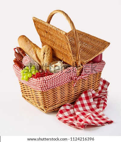 Packed picnic summer lunch in a wicker basket with fresh fruit, cheese, fruit and spicy sausages on a checked red and white cloth over white