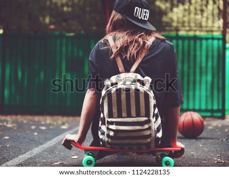 Young girl sitting on plastic orange penny shortboard on asphalt in cap
