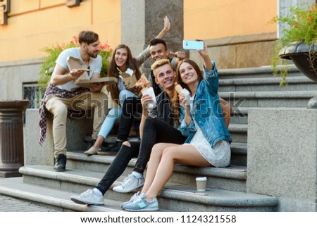 Selfie with best friends. Beautiful girls and boys sitting on stairs and eating hotdogs and pizza bought from a street vendor.