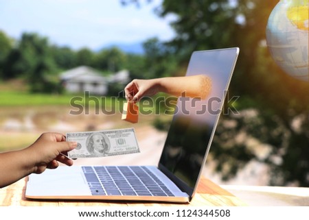 Hand holding shopping paper bag came out from laptop screen on wooden table outdoor and another hand holding one hundred dollar with nature background and globe. Online shopping and ecommerce concept.