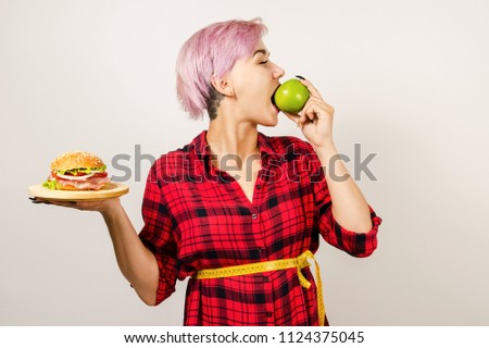 Young beautiful girl coiled measuring tape holds hamburger and eats green apple ,   on a light background.