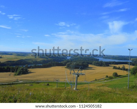 Large lake, ski lift, ski ropes, and a small lamp seen from the top of a high hill with a yellow pastureland and patches of trees seen at the bottom and visible horizon