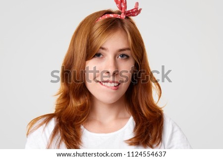 Happy satisfied female bites lower lips, has appealing look, long hair, wears headband, dressed in retro style, poses alone against white background. People, emotions and facial expressions concept
