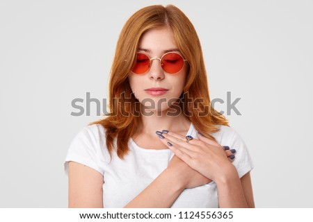 People and calmness concept. Pretty young female in stylish sunglasses, keeps eyes closed and hands on heart, touched by good words addressed to her, isolated over white wall, enjoys calm atmosphere