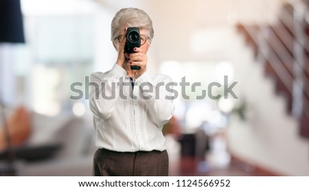 Beautiful senior woman excited and entertained, looking through a film camera, looking for an interesting shot, recording a movie, executive producer at home.