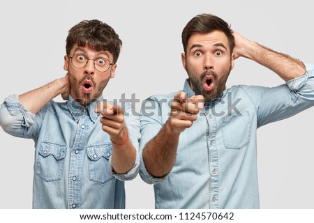 Terrific men point at camera as notices something attractive into distance, wears fashionable denim shirts, look alike, have stupefied expressions, isolated over white background. Reaction concept Royalty-Free Stock Photo #1124570642