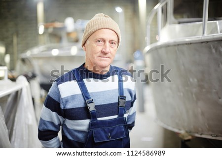 Mature engineer in workwear standing by new motor-boat or ship in workshop