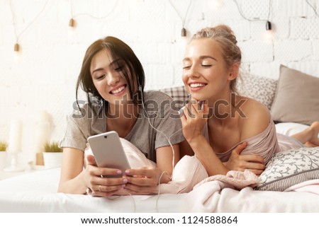 Portrait of joyful best friends listening music or viewing interesting film. Happy smiling young women lying in bedroom. Friendship concept. Blurred background