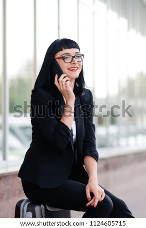 Young pretty girl dressed in a business suit and in the glasses with a suitcase talking on the phone and photographed