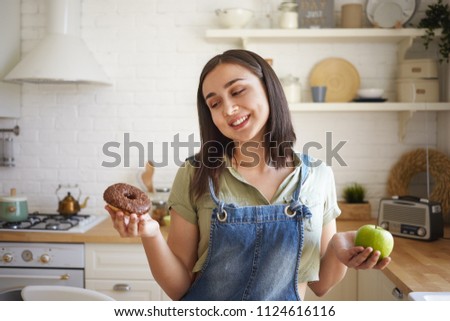 Health, food, nutrition, dieting and calories concept. Picture of smiling indecisive young chubby woman holding chocolate doughnut in one hand and green apple in other, feeling doubtful while choosing