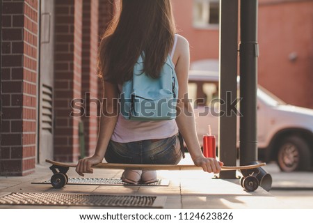 A girl with a blue backpack is sitting on a longboard in the evening. On the long board is a cocktail of cola.