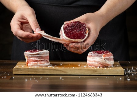Cooking beef meat with bacon stripes on wooden table by cook hands, copy text black background.