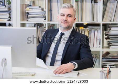 Portrait of mature business man with grey hair in his modern office with computer and many file folders on shelves on background