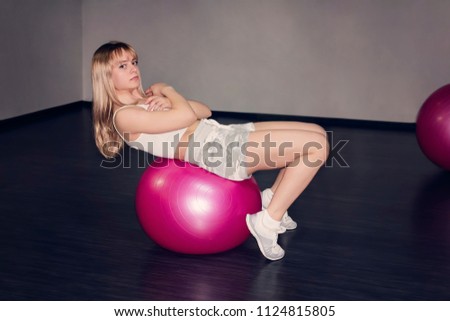 Blonde girl or young woman with green eyes and red lips lying on red pink gymnastic ball looking exhausted, tired, bored and weary wearing with training apparatus on background