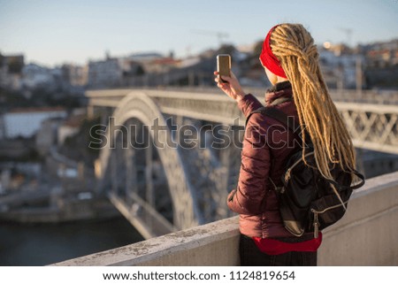 Young woman with dreadlocks takes pictures on a smartphone from the viewing platform opposite the Dom Luis I bridge in Porto, Portugal. 