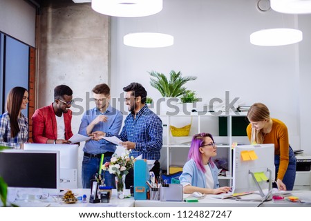 Well-lit white open office space with white furniture computer tables with digital gadgets. Multiracial Emploee People gathering near female manager with open laptop. Concept of a busy office life. Royalty-Free Stock Photo #1124827247