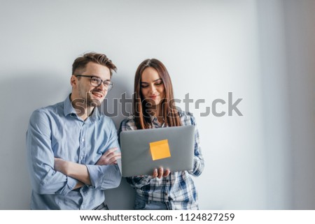 Portrait of two heterosexual colleagues in office interior, sharing useful information using laptop computers while standing isolated over gray wall background