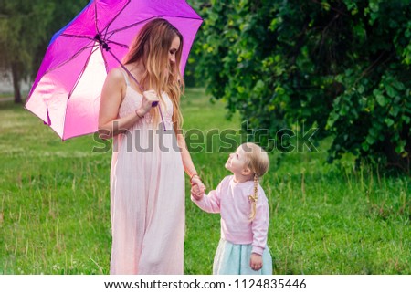 little and sweet blonde girl in a cute gray dress together with toothy smile beautiful mother walking under the summer rain with an umbrella in the park against the background of trees and greenery