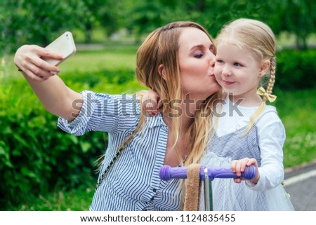 little and sweet blonde girl in a cute gray dress make selfie on phone together with beautiful mother have a rest in the park against the background of trees and greenery