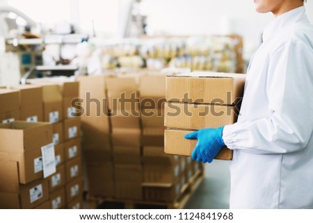 Young female worker in sterile cloths and blue gloves is carrying a stack of boxes to cargo room.