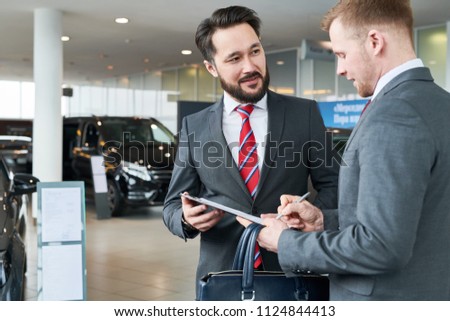 Businessman standing at auto salon with salesman. He is signing a contract