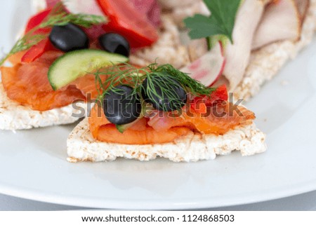 Sandwich with smoked salmon served on a rice waffle decorated with olive, cucumber, paprika and a sprig of fennel on a white plate closeup