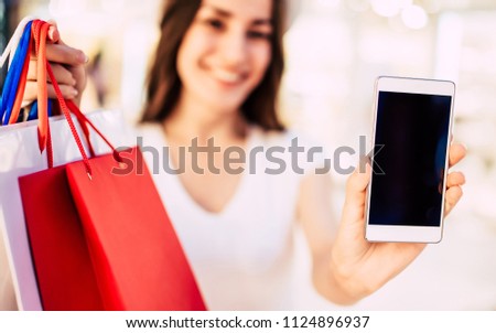 Close up photo of beautiful young woman in casual wear with shopping bags showing blank phone screen in hand on shopping center background. Cheerful buyer. Online Shopping