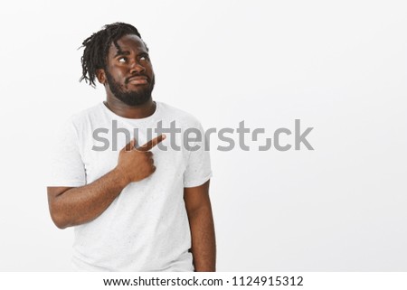 Guy are not sure about strange copy space. Portrait of intense doubtful plump african-american man with beard, looking and pointing at upper right corner, frowning and being unsure over white wall