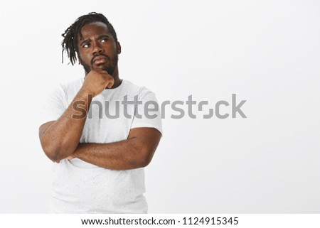 Choices and opportunities everywhere. Portrait of focused thoughtful young and plump african-american, touching chin with hand and staring frowned at upper right corner while thinking over gray wall