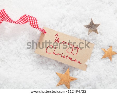 Gift tag with Merry Christmas greeting and a decorative red and white checked ribbon on a bed of winter snow with stars