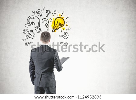Rear view of a young businessman in a gray suit holding a folder. He is looking forward. Question marks and a light bulb on a concrete wall. Mock up