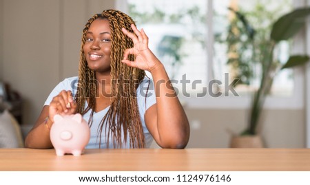 African american woman saving money with piggy bank doing ok sign with fingers, excellent symbol