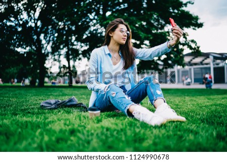 Young woman in spectacles posing for selfie on smartphone camera sitting in park during free time,hipster girl taking photo on mobile phone for blogging recreating on free time in college campus